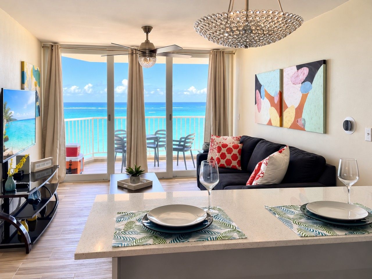 A modern living room with a balcony overlooking the ocean, a dining area with a table set for two, and a television on a stand.