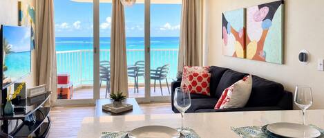 A modern living room with a balcony overlooking the ocean, a dining area with a table set for two, and a television on a stand.