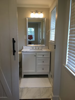 A narrow bathroom with a white vanity, a large mirror, and a window with blinds.