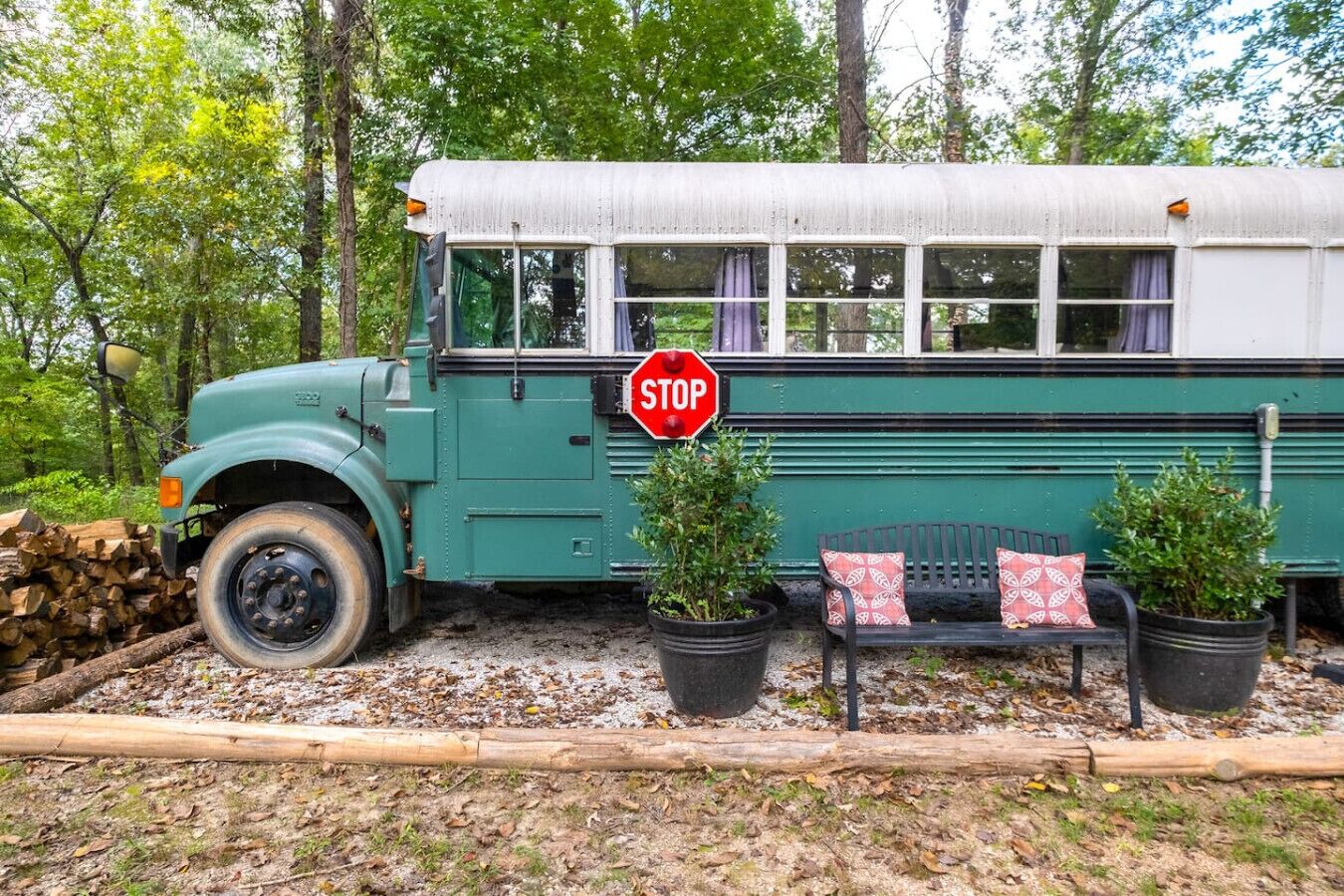 A green bus parked in a wooded area with a stop sign, a bench, and potted plants.