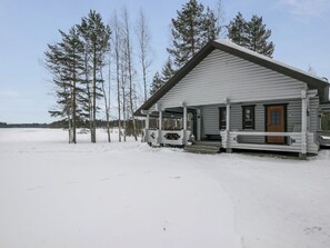 En træhytte med veranda og hoveddør, omgivet af sne og træer.