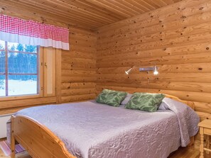 Et soveværelse i en træhytte med en seng, et vindue med et ternet gardin og et natbord.