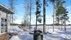 A modern house with a wooden deck and a large outdoor fireplace surrounded by snow and trees. - Suvituuli by Interhome (Pertunmaa)