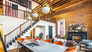 A modern living room with a large island table, orange chairs, a fireplace, and a staircase with a wooden railing. - Historic DC Firehouse Penthouse Loft (Washington)