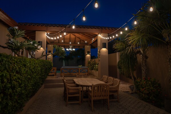 An outdoor patio area with a wooden table and chairs, string lights, and palm trees.
