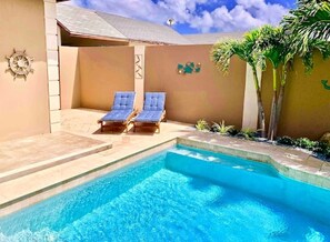 Une piscine avec deux chaises longues, un palmier et un mur orné d’une boussole et d’une horloge.