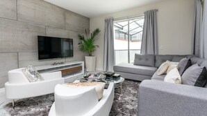 Een moderne woonkamer met een flatscreen televisie, grijze banken, een salontafel en een groot raam met gordijnen.