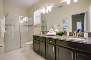A modern bathroom with a glass-enclosed shower, dark cabinets, a large mirror, and a granite countertop. - Tempo Lane Villa Champions Gate (Davenport)
