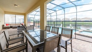 A spacious patio area with a glass-top table and chairs, a cushioned seating area, and a view of a swimming pool and buildings in the distance.
