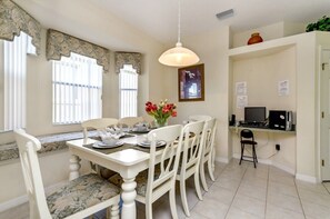 Un comedor con una mesa blanca dispuesta para cuatro personas, un jarrón de flores rojas y una computadora en un escritorio.