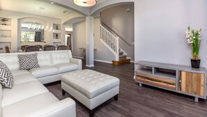 A spacious living room with a white sofa, a large ottoman, and a grey entertainment unit. A staircase with a white railing leads to an upper level. - Oasis Key Villa Emerald Island (Kissimmee)