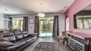 A modern living room with a leather sofa, a dining table, and a large window with curtains. - Oasis Key Villa Emerald Island (Kissimmee)