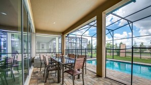 Une terrasse couverte avec une table et des chaises en verre, une piscine en arrière-plan et une vue sur l’extérieur.