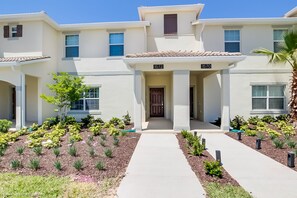 A two-story building with a central entrance, numbered 1570, surrounded by a landscaped garden with various plants and a paved walkway. - Stinger Drive Villa Champions Gate 2 (Davenport)