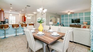 A modern kitchen with bar stools, a dining table set for six, and a living area with a sofa and television. - Showhome 5 Bedroom Champions Gate Villa 1401mvd (Davenport)
