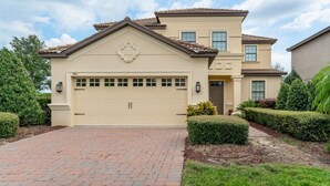 A two-story house with a beige facade, a garage, and a brick driveway. - Showhome 5 Bedroom Champions Gate Villa 1401mvd (Davenport)