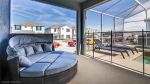 A room with a large window, a wicker sofa, and a view of a swimming pool and neighboring houses.