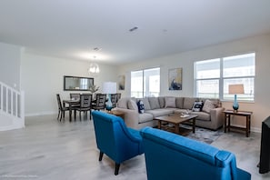 A spacious living room with a large sectional sofa, two blue armchairs, a coffee table, and a dining area with a chandelier.