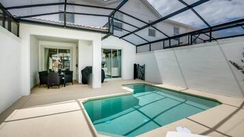 A covered swimming pool with a glass ceiling, a dining area with chairs and a table, and a white exterior wall.