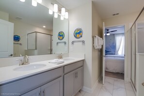 A bathroom with two sinks, a large mirror, and a shower. There is a towel rack and a door leading to a bedroom with a bed and a window. - 8911sd Champions Gate 8 Bedroom Resort Villa (Davenport)