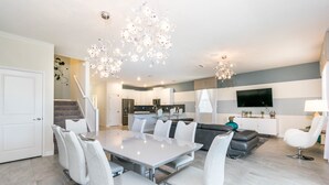 A modern kitchen with a built-in oven, a dining area with a white table and chairs, and a living area with a gray sofa and a TV mounted on the wall.