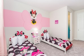 Un dormitorio con dos camas, cada una con una colcha temática de Minnie Mouse. Hay una mesita de noche con una lámpara y un peluche de Mickey Mouse. La habitación tiene paredes blancas y una pared de acento rosa con una silueta grande de Minnie Mouse.