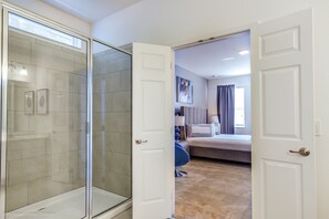 A hotel room with a glass shower enclosure, a bed with a headboard, a bedside table, and a large window with curtains. - 7013ows 14 Bed 11 Bathroom (Davenport)