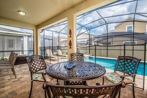 An outdoor patio with a round table and chairs, a pool, and a view of a house through a screened area. - Amazing 6 Bedroom 5 Bathroom Solara Resort Villa (Kissimmee)