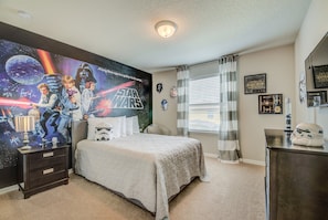 Une chambre avec une fresque murale sur le thème de Star Wars, un lit recouvert d’une courtepointe, une table de chevet, une commode et une fenêtre avec des rideaux.