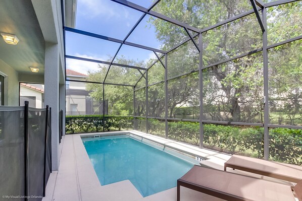 A swimming pool with a glass enclosure, surrounded by greenery and a view of a house.