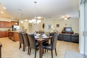 A dining area with a wooden table and chairs, a kitchen in the background, and a living room with a sofa and television.