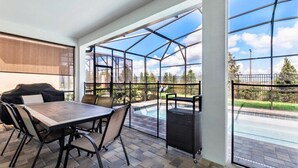 A covered patio area with a dining table and chairs, a grill, and a view of a swimming pool and greenery.