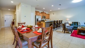 A spacious dining area with a wooden table set for four, a kitchen in the background, and a living room with a sofa and coffee table. - Cuban Place 8963 Paradise Plams (Kissimmee)