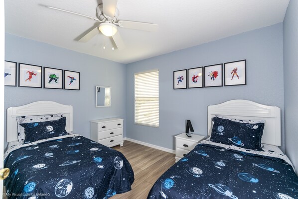 A bedroom with two beds, a ceiling fan, framed superhero posters, a window with blinds, and a nightstand with a lamp.