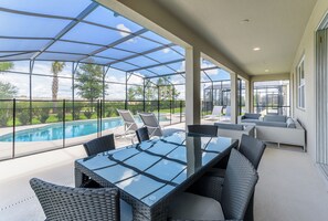 Un espacio al aire libre techado con una mesa y sillas de vidrio, una piscina y un área de estar con sofás.