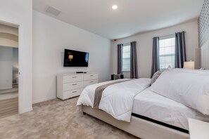 Un dormitorio con cama, televisor sobre una cómoda, ventana con cortinas y puerta que da a otra habitación.