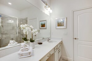 A modern bathroom with a large mirror, white countertop, and a sink. There are towels stacked on the counter, and a vase with white flowers. - Oakgrain Villa Solterra 1 (Davenport)