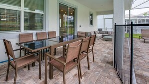 Ein überdachter Patio-Bereich mit einem Esstisch und Stühlen aus Glas, einer Couch und einer Glastür, die in einen Garten führt.
