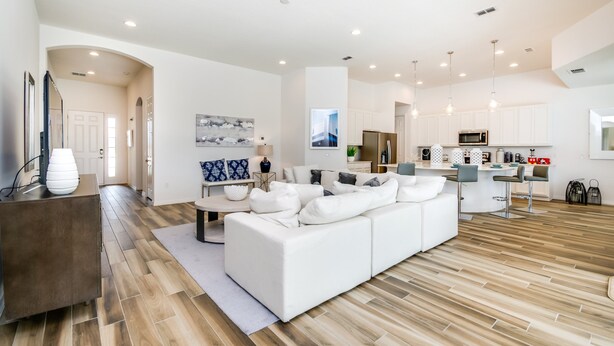 A modern living room with a white sofa, a coffee table, and a TV on a wooden cabinet. There is a kitchen area with white cabinets and a microwave.