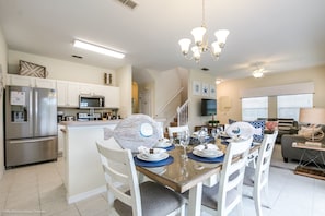 Una cocina moderna con gabinetes blancos, electrodomésticos de acero inoxidable y un comedor con mesa de vidrio y sillas blancas.