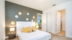 A bedroom with a white bed, beige carpet, and a wall with hexagonal light fixtures.