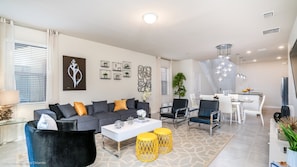 A modern living room with a grey sofa, yellow ottomans, and a white coffee table. There is a dining area with a white table and chairs, and a kitchen in the background.
