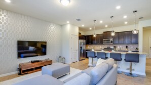 A modern living room with a large flat-screen TV, grey sofas, and a kitchen area with dark cabinets and a bar stool seating. - 4659terra (Davenport)