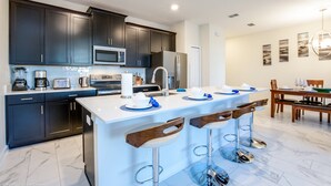 A modern kitchen with dark cabinets, a central island with bar stools, a dining area with a table and chairs, and a large window providing natural light. - Stinger Drive villa 8953 Champions Gate (Davenport)