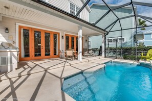 A pool area with a covered patio, outdoor seating, and a view of a house with a screened porch.