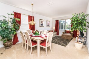 Un coin repas avec une table dressée pour quatre personnes, entourée de chaises, et une table à manger agrémentée d’un chemin de table rouge. Deux plantes en pot sont disposées de part et d’autre de la table.