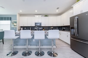 A modern kitchen with white cabinets, a stainless steel refrigerator, and three white bar stools. - Flange Drive Champions Gate 1564 (Davenport)