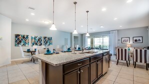 A modern kitchen with a large island, pendant lights, and a dining area with striped chairs. - 8887menton Ultimate 8 Bedroom 6 Bathroom Windsor (Kissimmee)