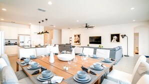 Una cocina moderna con armarios blancos, un comedor con una mesa larga preparada para seis comensales, y una sala de estar con un sofá gris y un televisor en la pared.