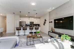 Una sala de estar moderna con un sofá, una mesita de centro y un televisor. Se ve una cocina al fondo, con un refrigerador, microondas y taburetes de bar.
